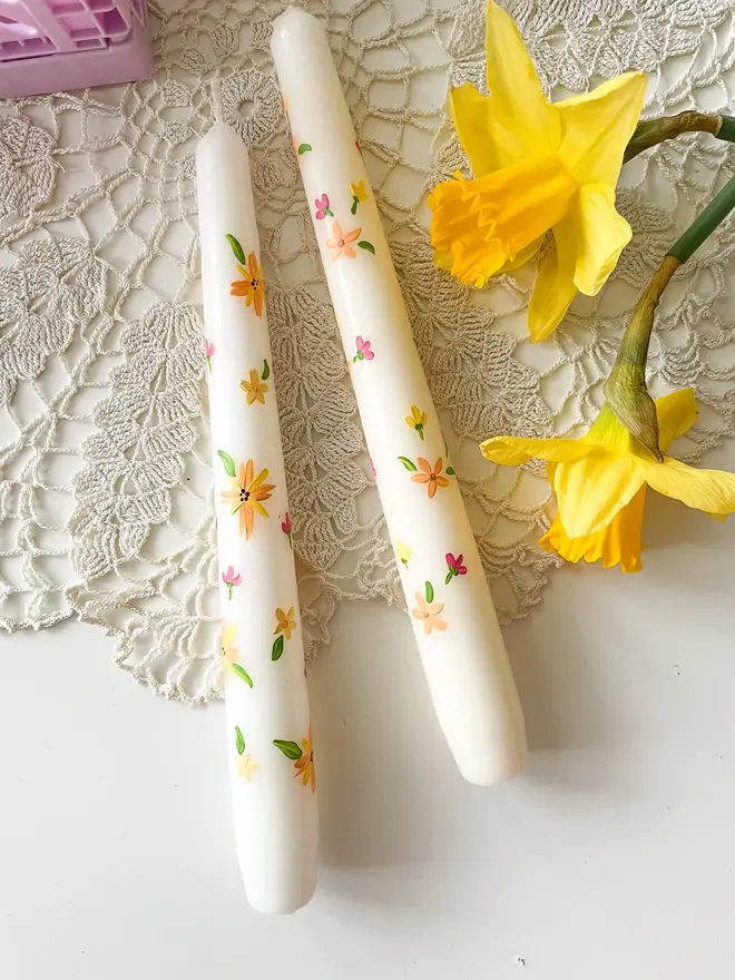 Blooming For You & Spring Bound Hand Painted Candle, a pair of taper candles against a white desk. The floral design can be seen wrapping around them. They are accompanied by a flower.