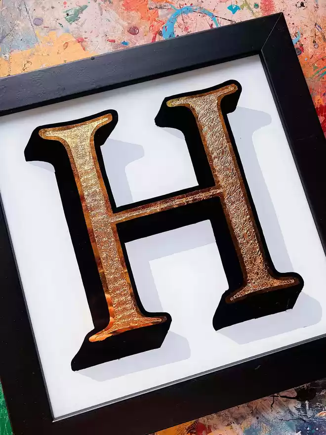 Hand-painted and gilded gold leaf 'H' with painted white background in a black frame, on a painty desk.