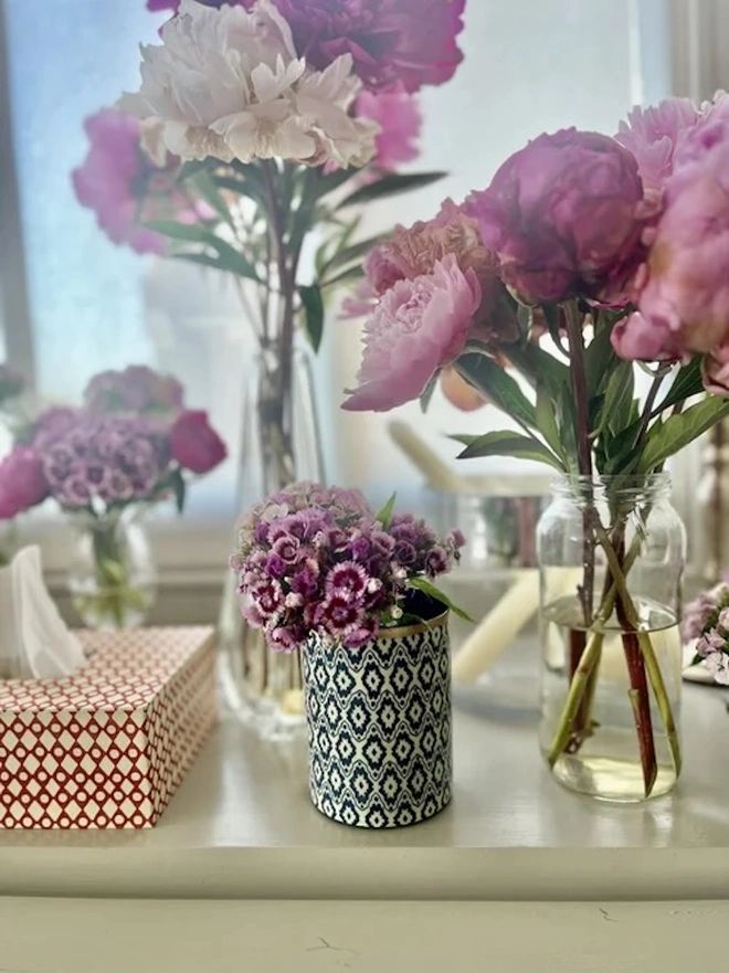 Perfect Enamel Pot. A navy and cream ikat-patterned pot with a gold rim filled with purple sweet williams, styled on a windowsill beside peonies in glass vases. A matching red diamond-print tissue box completes this colourful home decor display.