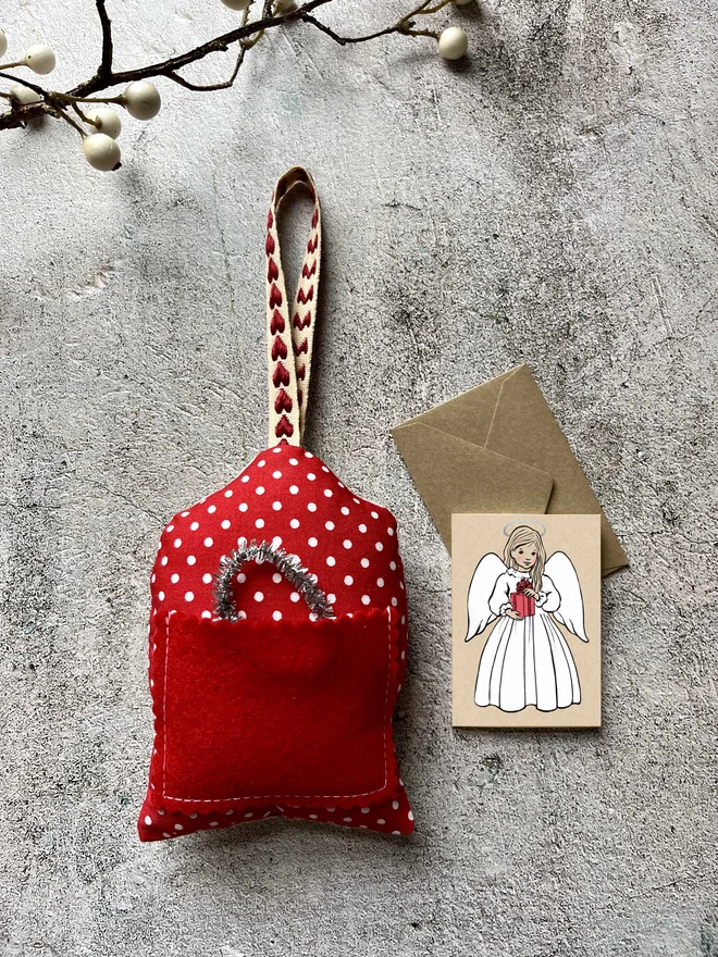aan image showing the back of a christmas tree hanging angel decoration with a red pocket with a tinsel halo in it and next to it is a mini greeting card resting on a kraft envelope the greeting card shows an angel holding a pink present