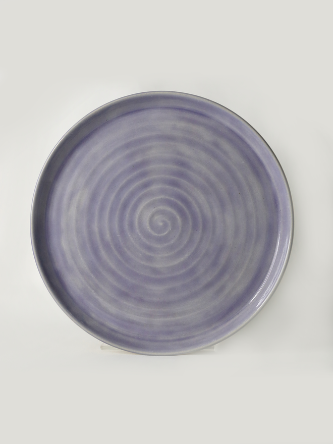 A handmade ceramic lunch plate in glossy amethyst purple glaze, photographed from above against a plain light grey background, showing a natural concentric swirl texture from the wheel-throwing process