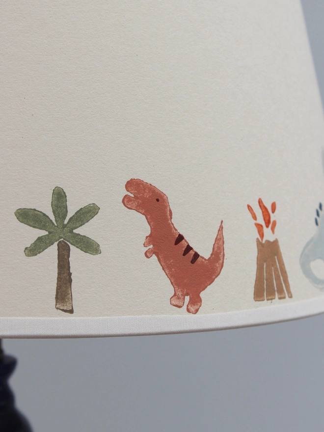 Close-up of a Studio Zef children's dinosaur lampshade, featuring hand-printed illustrations of a terracotta red T-Rex, green palm tree, erupting volcano and pale blue dinosaur. The playful prehistoric motifs are printed along the lower edge of the cream card empire lampshade, showing the textured finish and charming imperfections of the traditional lino printing process.