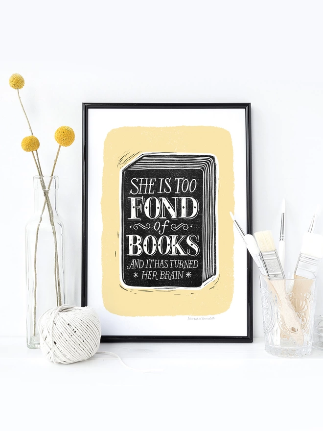 she is too fond of books black and yellow print in a black frame with yellow flowers, string and make up brushes