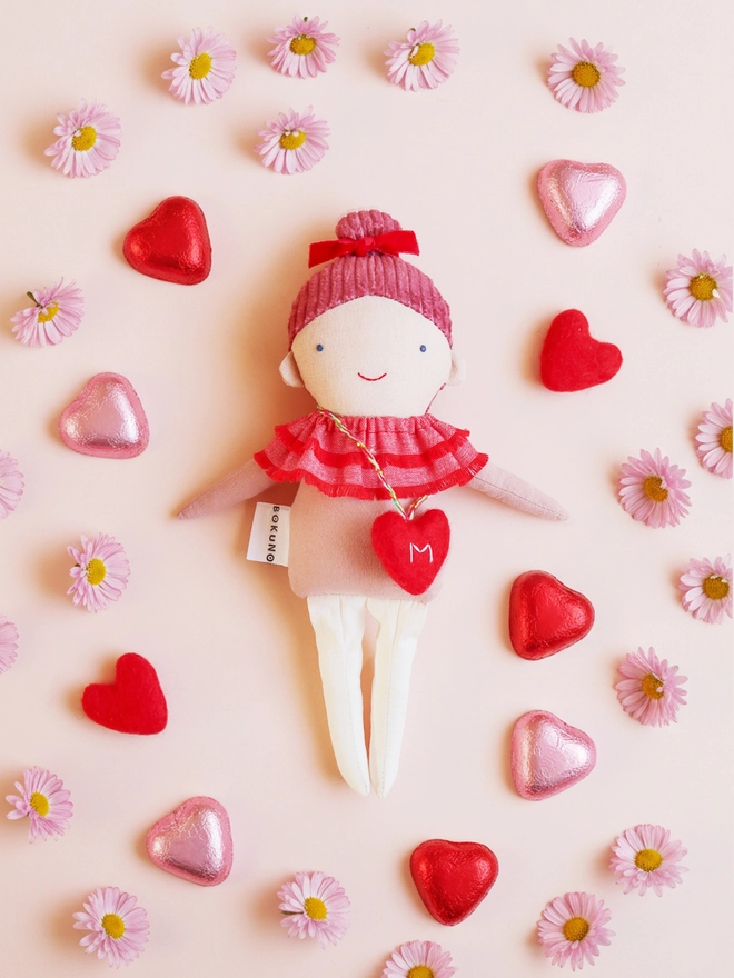 Handmade cloth doll with a pink corduroy hair with a red velvet bow and a layered red ruffle collar and pink outfit, wearing a small red felt heart bag with the letter “M”