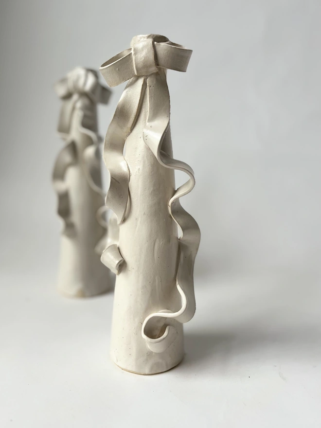 two cream white stoneware tall candlestick holders with a ceramic bow with long ribbons flowing down the body of the candlestick itself