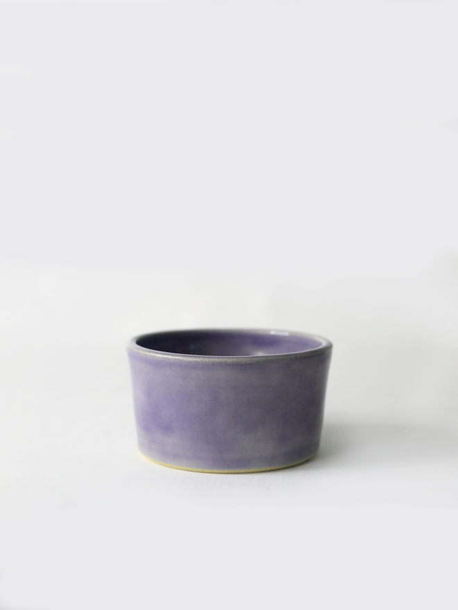A small handmade ceramic side bowl in glossy amethyst purple glaze, with a tapered sides and a wide open rim, photographed against a plain light grey background