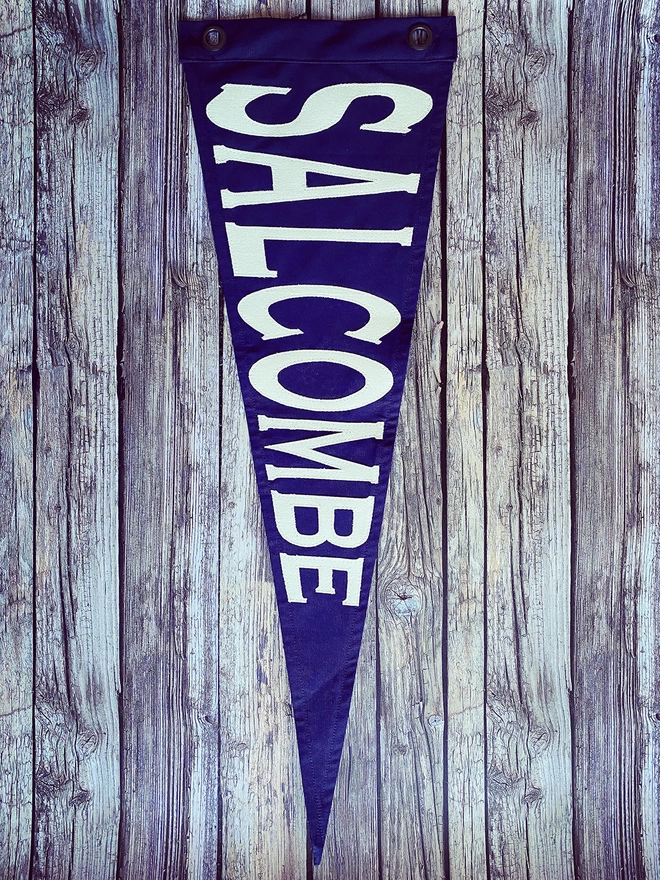 A navy pennant with the place name Salcombe on ivory. Its placed on a wooden wall