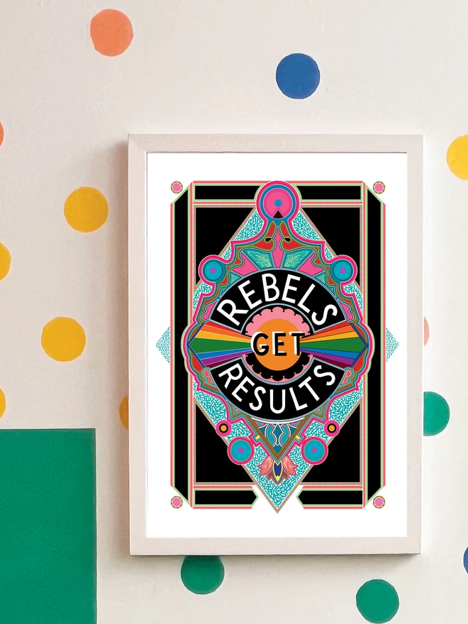 Rebels Get Results is written in white on a black background at the centre of this vibrant, abstract portrait illustration, with a black background and rainbows emitting from the centre, and blue and pink detailing.. The picture is hanging in a white frame on a white wall, with yellow, orange, green and blue spots and a green and yellow rectangle painted in the top left hand corner.