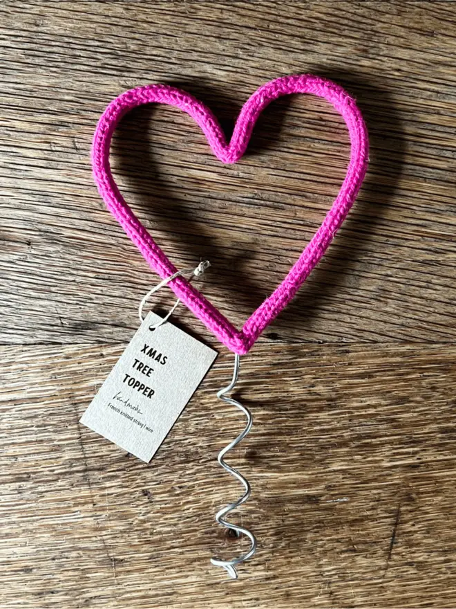 A pink string wrapped wire heart shaped Christmas tree topper on an oak table