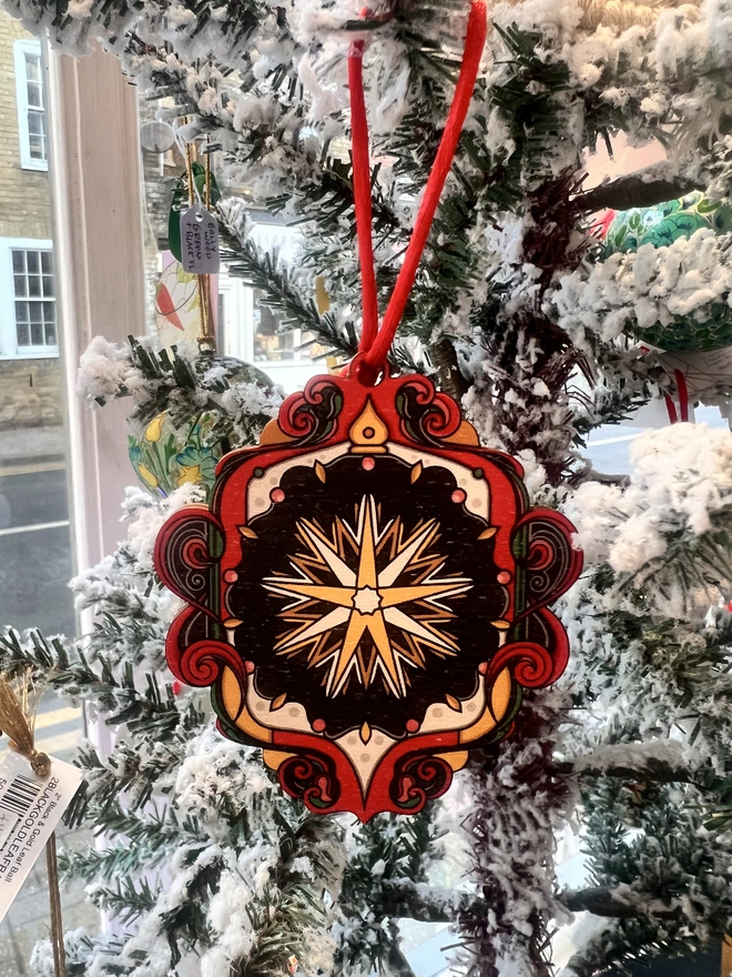Multi coloured wooden tree decoration with a star pattern at the centre. It is hanging by a red ribbon on a fake white christmas tree with other decorations. 