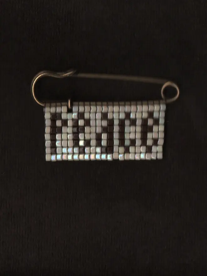 'lingua ii' beaded 'peace' pin. a beaded pin badge with the word peace in brown lettering on a pearl background.