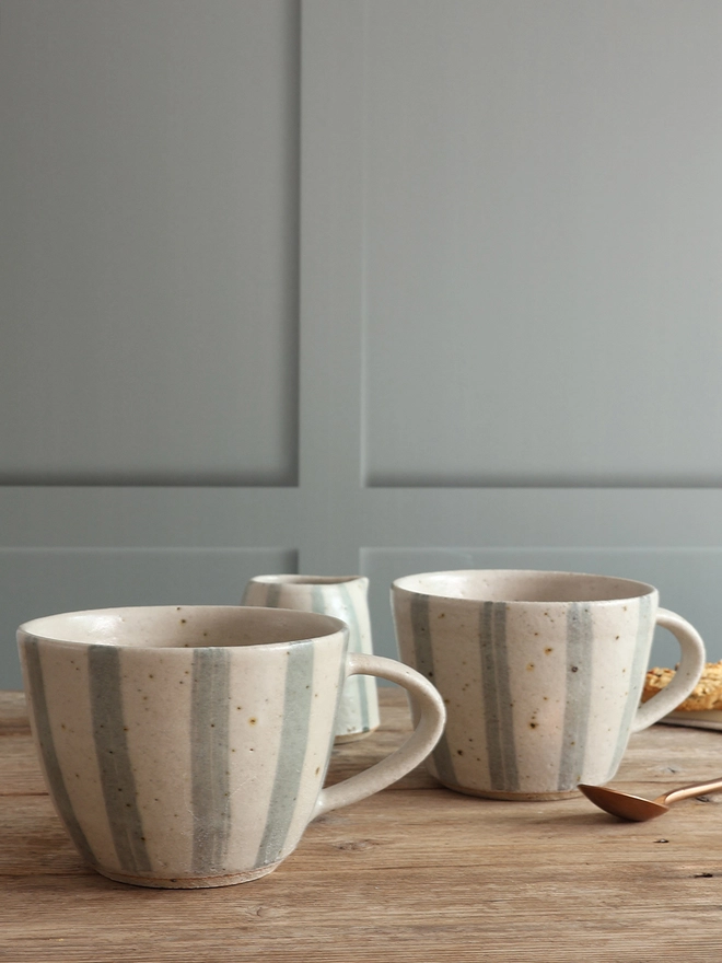 Handmade Ceramic Stoneware Mug in Blue and White Stripe sat on a wooden surface. The background shows another of the same mug sat alongside a handmade small jug in the same style. 