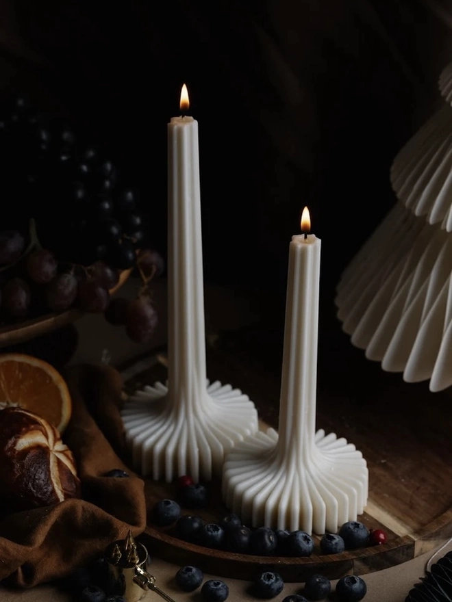 Pair Of White Flair Candles (Set Of 2). Two lit cream sculptural candles with slim fluted stems that flare into wide ribbed bases, on a wooden board with blueberries, grapes and orange slices. The self-standing design needs no holder, so they’re a striking choice for festive tables.