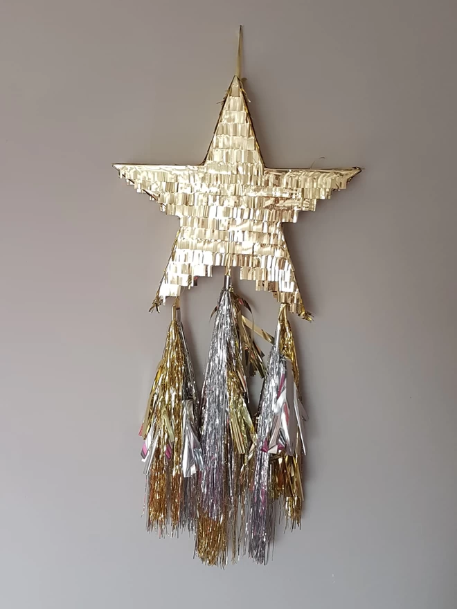 Gold shining star pinata with gold and silver tassel tails against a grey background