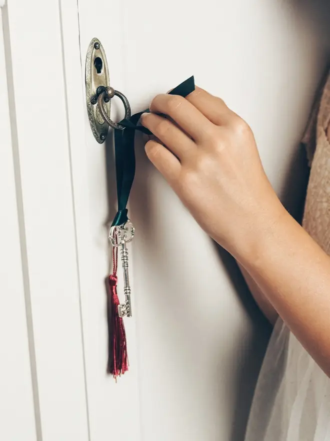 A child's hand ties the key's green satin ribbon to a brass door handle. The ornate silver key and red tassel hang below.