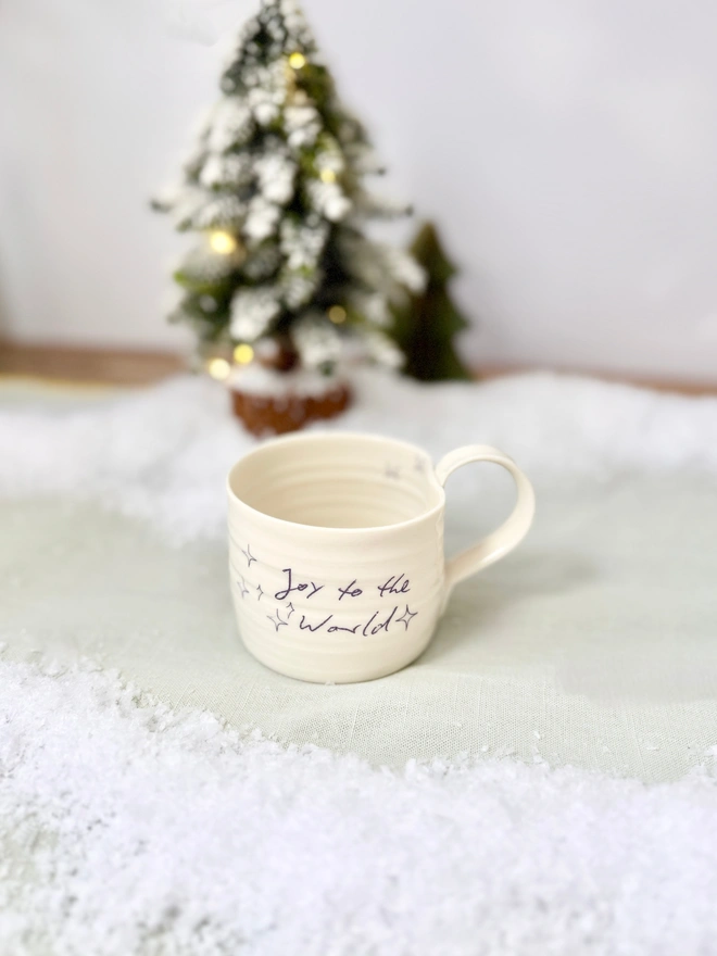 cream hand thrown porcelain mug with the words 'Joy to the World' printed onto the side with pretty line drawn stars. Sitting on a tabletop with snow, a mini lit Christmas tree 