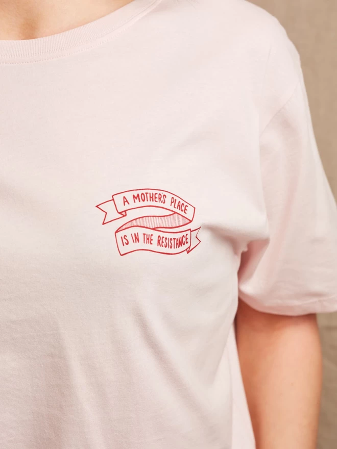  Close up of the front of the pink t-shirt showing the words A Mother’s Place is in the resistance written in red letters on a ribbon, on the left breast 