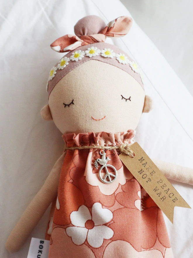 Handmade fabric doll called Peace Wishing Flower Child Doll, wearing peach and white retro floral dresses, daisy headbands, and bow topknots. It has serene embroidered face, peace sign charm around its neck, and tags reading “Make Peace Not War.” 