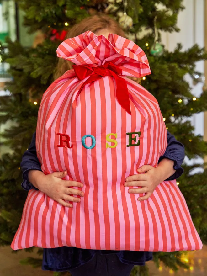 Small child holding a personalised striped Christmas sack spelling out 'ROSE' in front of a Christmas Tree