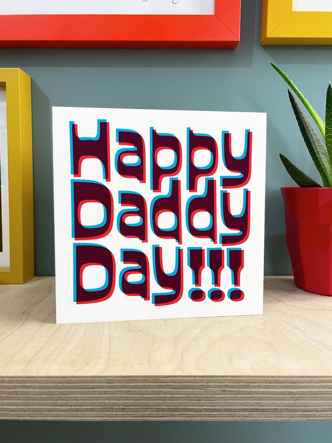 Happy Daddy Day printed in magenta and cyan blue ink, a slight overlapped design to create this retro jolly card. Stood on a ply shelf with a plant to one side and coloured frames around.