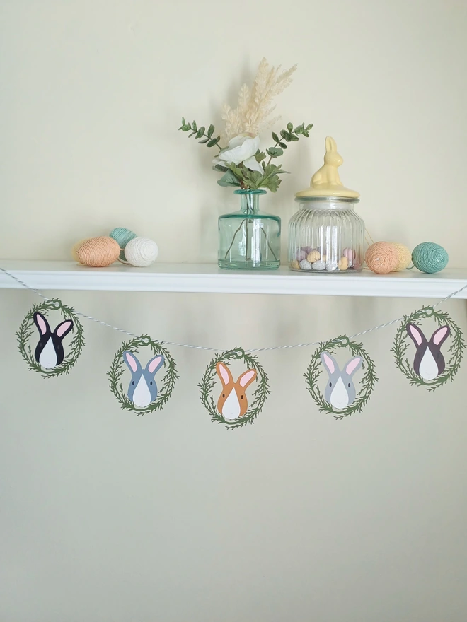 Easter Garland with 5 Decorations hugn from shelf. Decoration is a green card lasercut foliage wreath framing a bunny face. Black, Dark grey, Toffee, Light Grey, Dark Brown rabbits each with pink ears and white nose