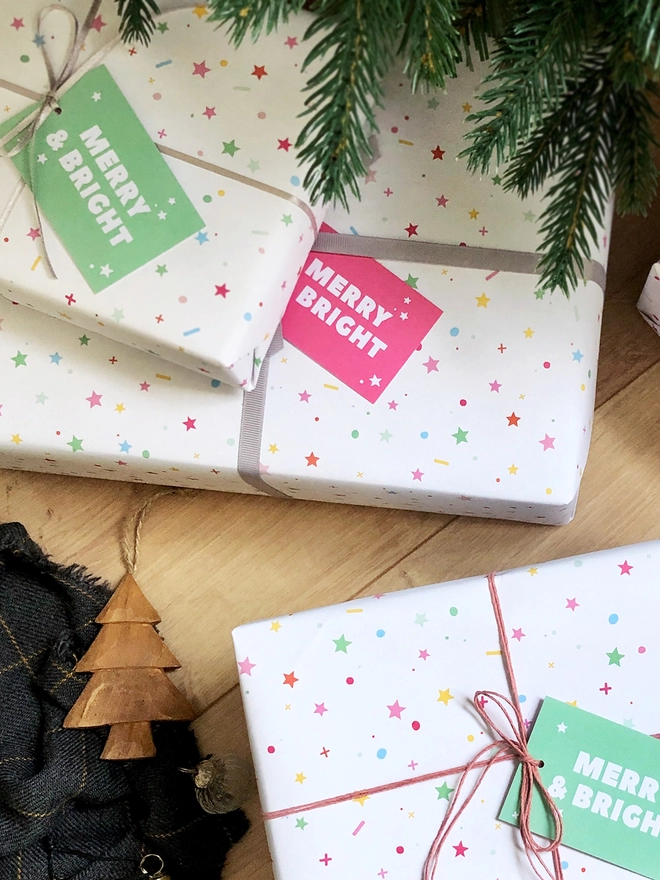 Gifts wrapped in pastel star Christmas wrapping paper are on a wooden floor beneath a Christmas Tree.