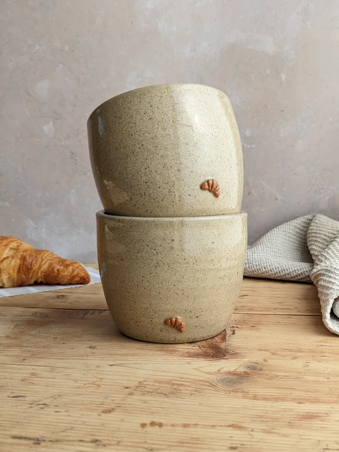 Two croissant coffee cups in biscuit glaze stacked on top of each other against a grey background.