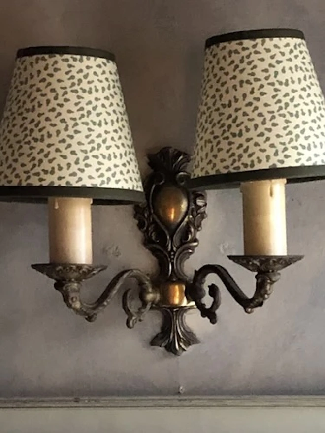 Candle Clip Shades. A pair of small candle shades in a cream and green scattered leaf print with dark trims, fitted to an ornate antique twin-arm wall sconce. The patterned shades add a refined, traditional finish to period wall lighting.
