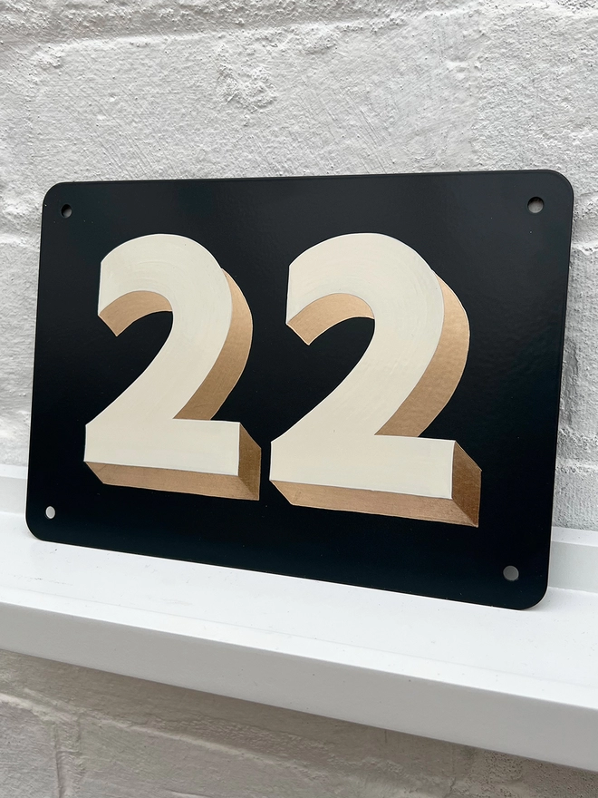 Hand painted off white and gold leaf house number 22 on an anthracite grey metal plaque against a white brick wall. 