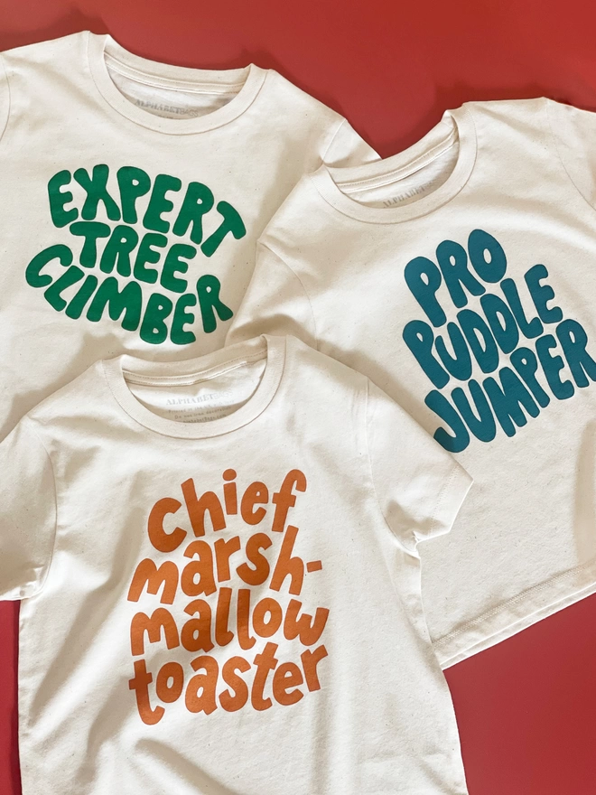 three slogan organic cotton t-shirts laying on an orange background. One reads Expert Tree Climber, the second reads Pro Puddle Jumper and the third reads Chief marshmallow toaster