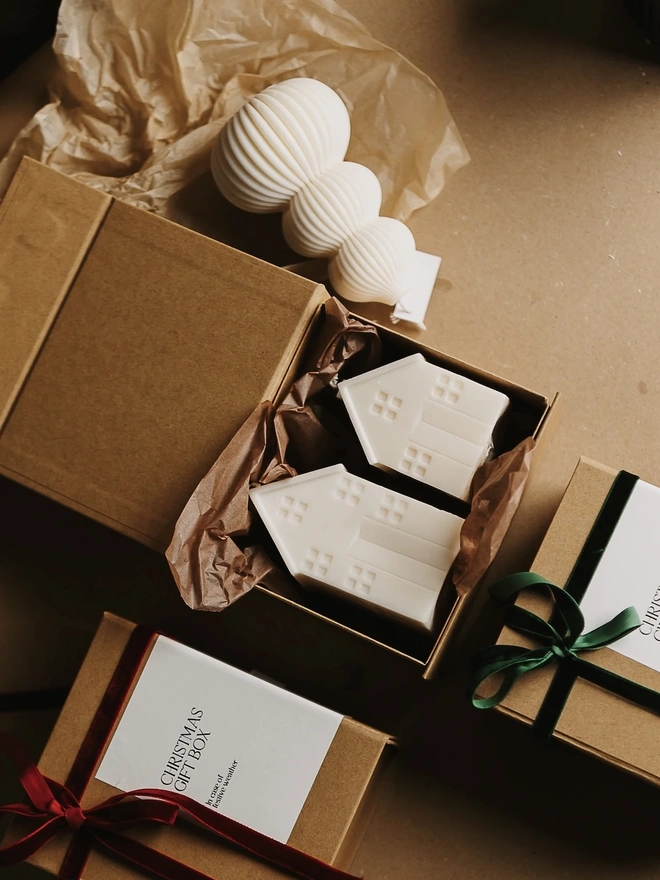 White Christmas Townhouse Candles (Set Of 3). A kraft Christmas gift box holding two cream house candles in brown tissue, beside boxes tied with red and green velvet ribbons and a ribbed tree candle. The gift-ready packaging makes festive giving simple and stylish.