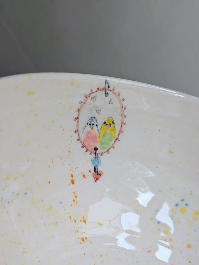 close up of a ceramic bowl with hand painted mirror and budgie couple the colourful splatters and hearts