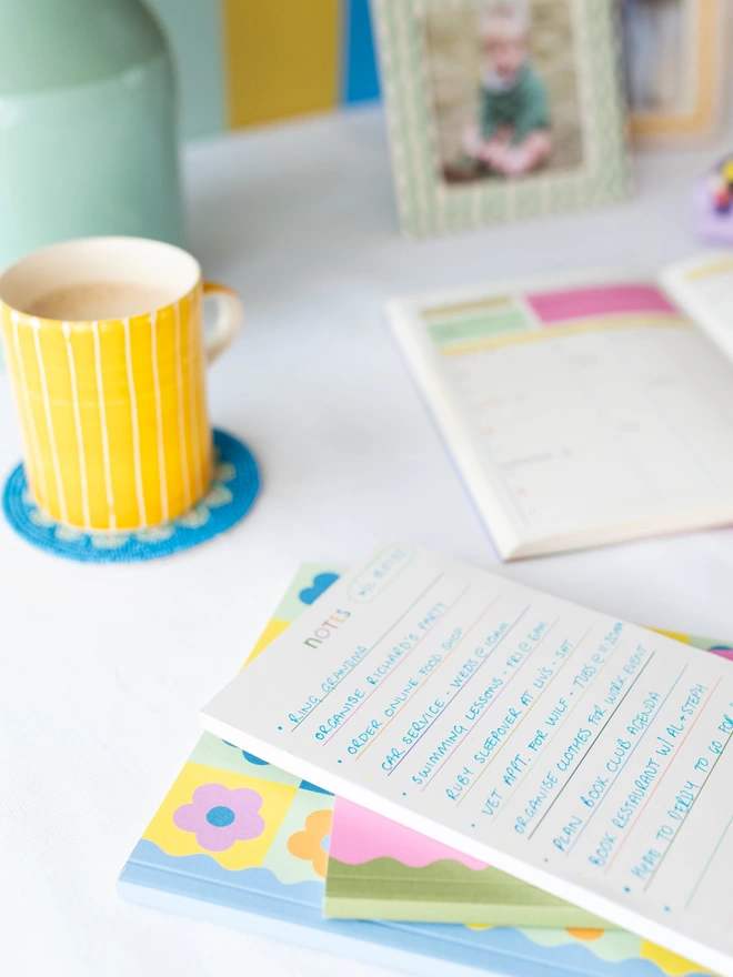 The Raspberry Blossom rainbow coloured lined note pad sits on a desk in a pile of other colourful stationery items from the Happiness collection