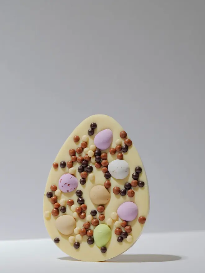 Large white chocolate egg-shaped tablet decorated with pastel mini eggs and assorted chocolate pearls, standing upright on a plain white background.