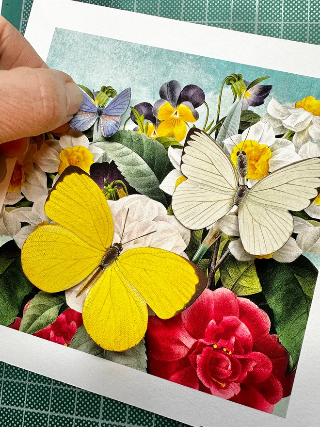 Hand placing a blue paper hand cut butterfly on a floral print with a yellow and white butterfly.