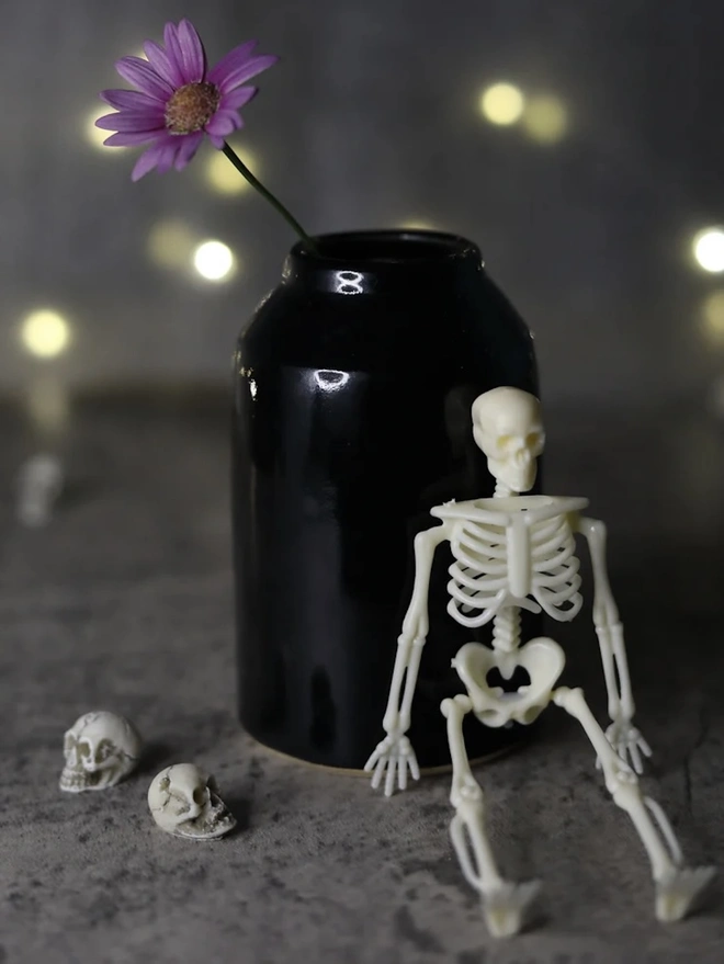 Classic Bud Vase, a colourful ceramic vase sitting on a plain surface against a grey backdrop. It is accompanied by some small skeletons and a flower. 