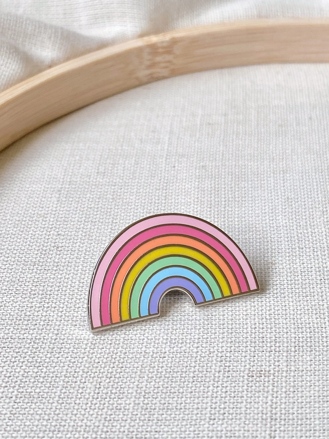 A small pastel rainbow enamel pin badge is laid on beige fabric.