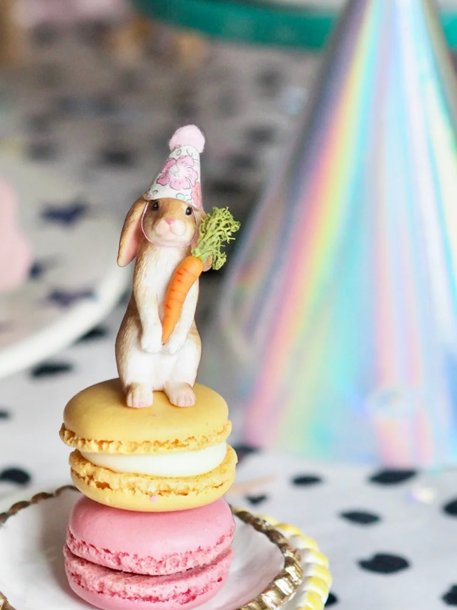 Rabbit with Carrot Cake Topper Decoration. A standing rabbit in a party hat and holding a carrot, set atop two macarons.