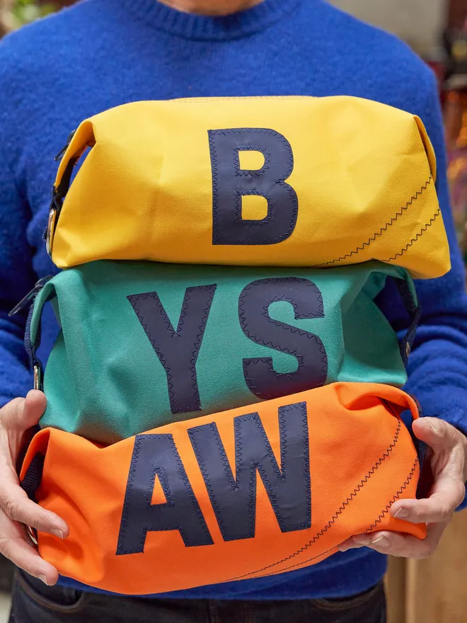 Personalised canvas wash bags in yellow with a 'B', green with a 'YS' and orange with an 'AW', held by a man in a blue jumper