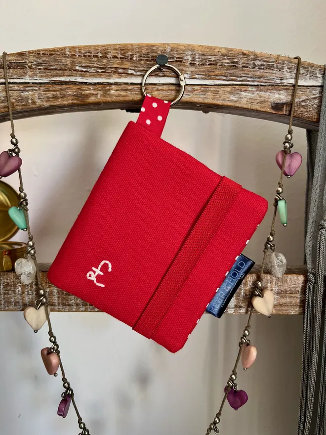 Red card wallet hanging by the attached silver hinged keyring. It has a red elastic closure and a £ sign embroidered in white thread on the front. 
