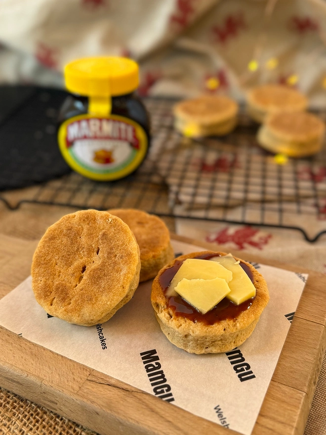 Marmite and Cheese Welsh Welsh cakes sitting on a wooden chopping board in a messy rustic kitchen with marmite spread and cheese on top of one welsh cake.