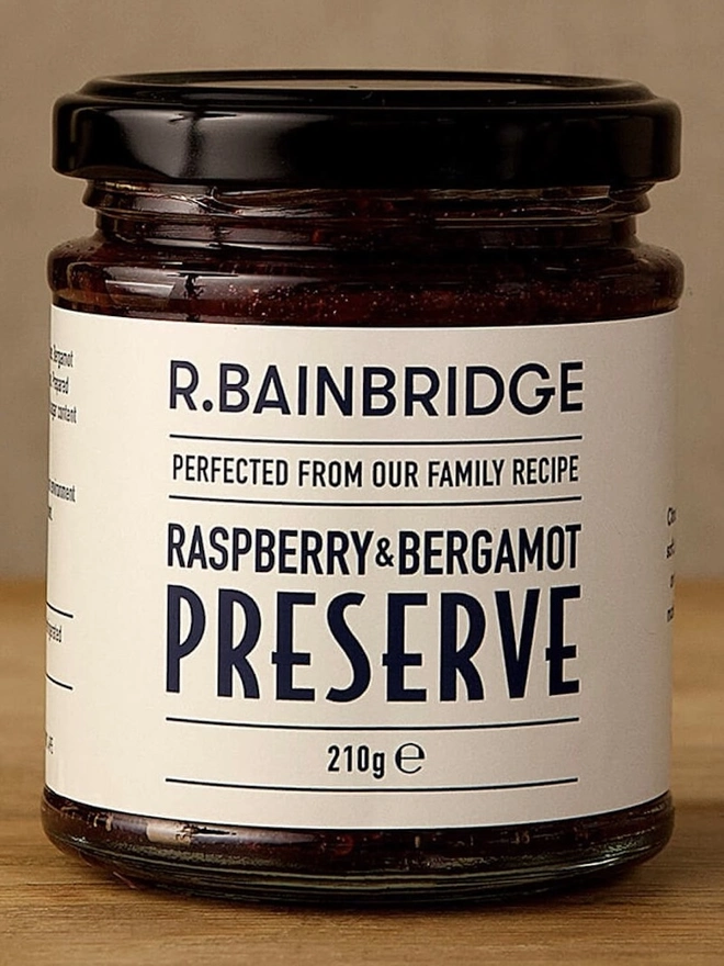 Raspberry & Bergamot Jam (Set Of 1, 2 Or 3), a jar of jam sitting on a wooden surface in front of a plain backdrop. 