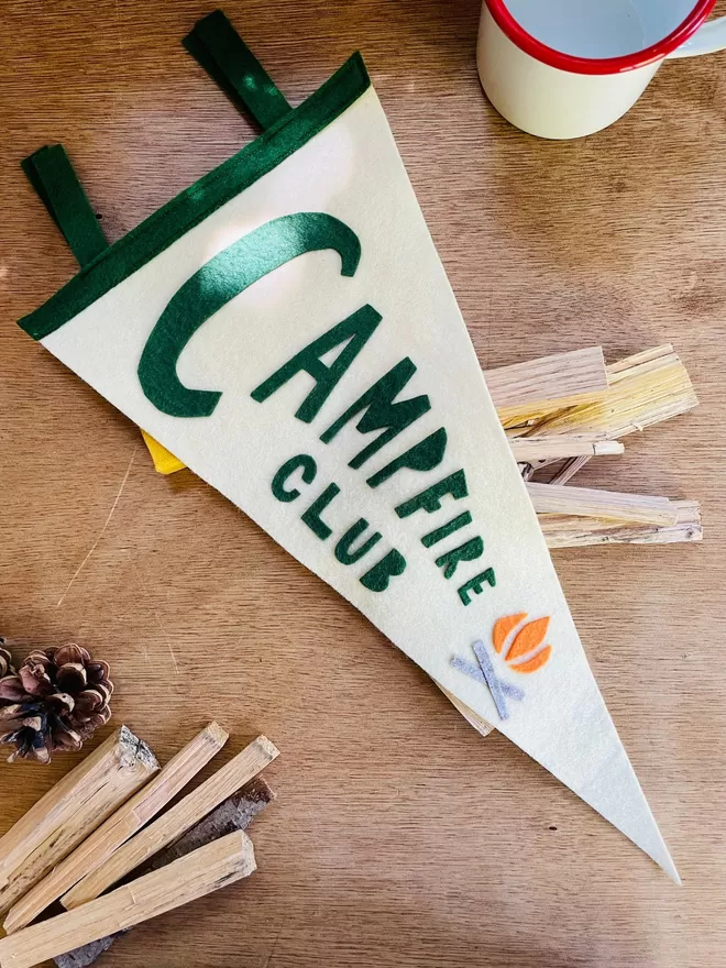 Campfire Club pennant flag laying on some firewood on a wooden table.