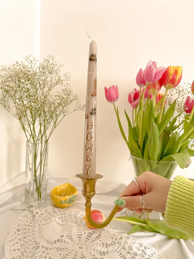 'Howdy' Personalised Hand Painted Candle, a woman placing the candle on a table top accompanied by flowers. It's design can be seen wrapping around the stick. 