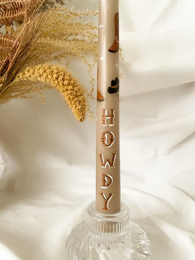 'Howdy' Personalised Hand Painted Candle, a candle in a glass holder on a white surface accompanied by a plant. It's design can be seen wrapping around the stick. 