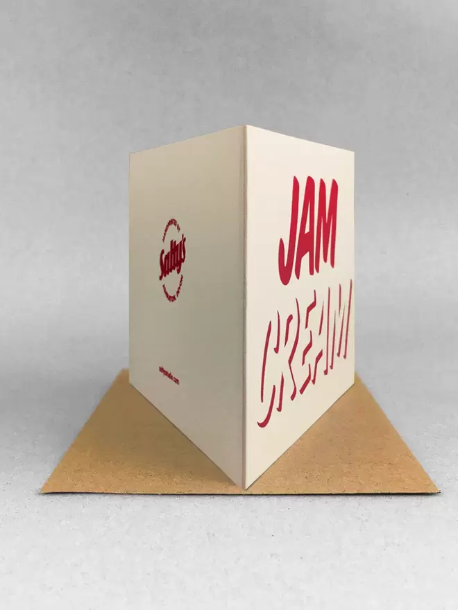 Rear view of a cream card with the words Cream at the bottom, Jam on top, printed in red ink. Stood on a kraft brown envelope in a light grey studio with soft shadows.
