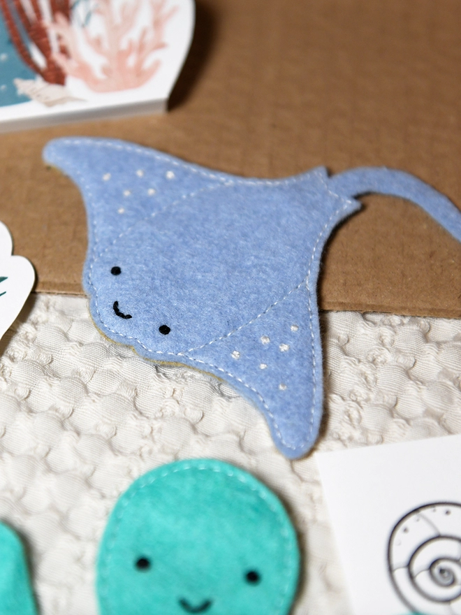 A soft blue felt manta ray has been sewn together and is on a wooden desk beside craft kit materials.
