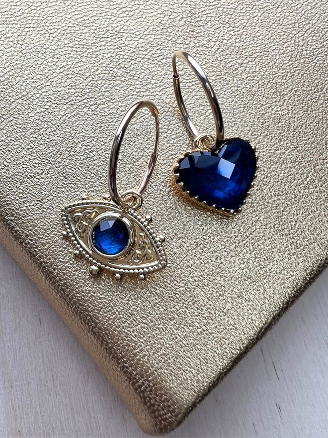 Mismatching gold hoop earrings with blue evil eye charm and blue faceted heart charm on gold background 