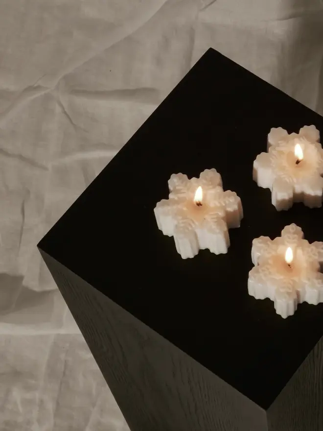 Classic Cream Snowflake Sculpture Candle. Three lit snowflake candles glowing on a black wooden plinth against a draped linen backdrop. The minimalist festive candles create a calm, elegant mood for winter evenings.