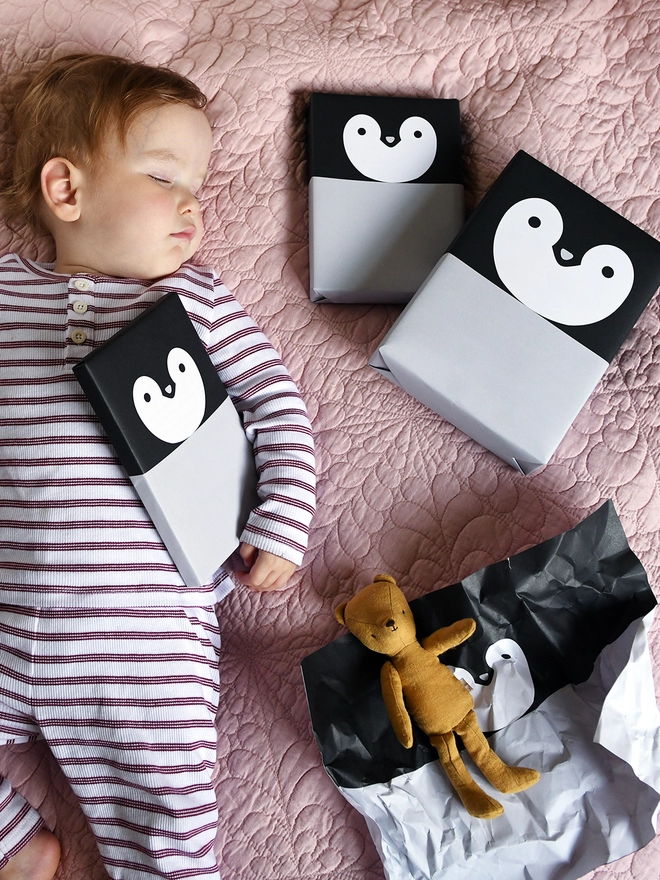 A baby wearing red and white striped pyjamas is asleep on a pink quilt with several gifts around them, all wrapped as penguins.
